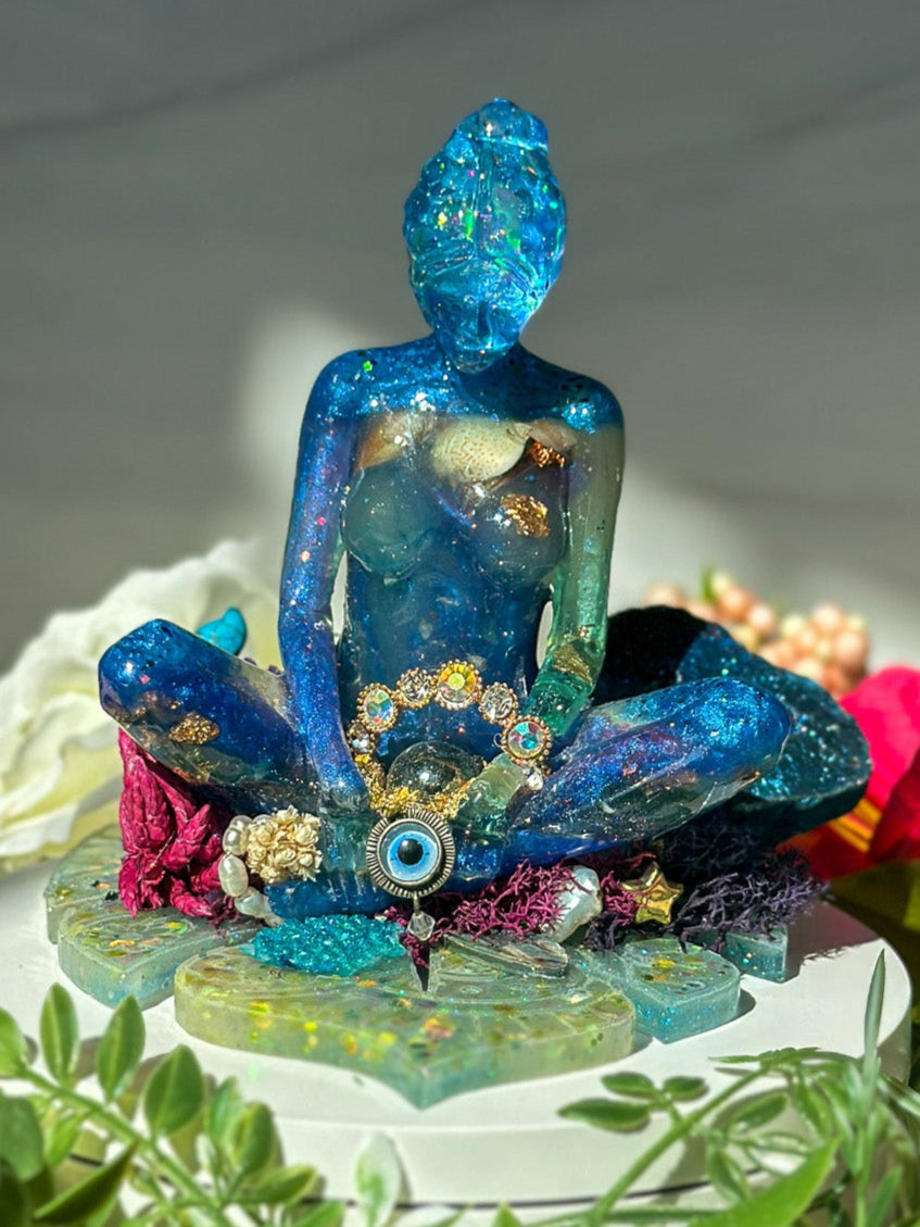 Colorful meditating sculpture statue on a white platform with flowers and greenery surrounding