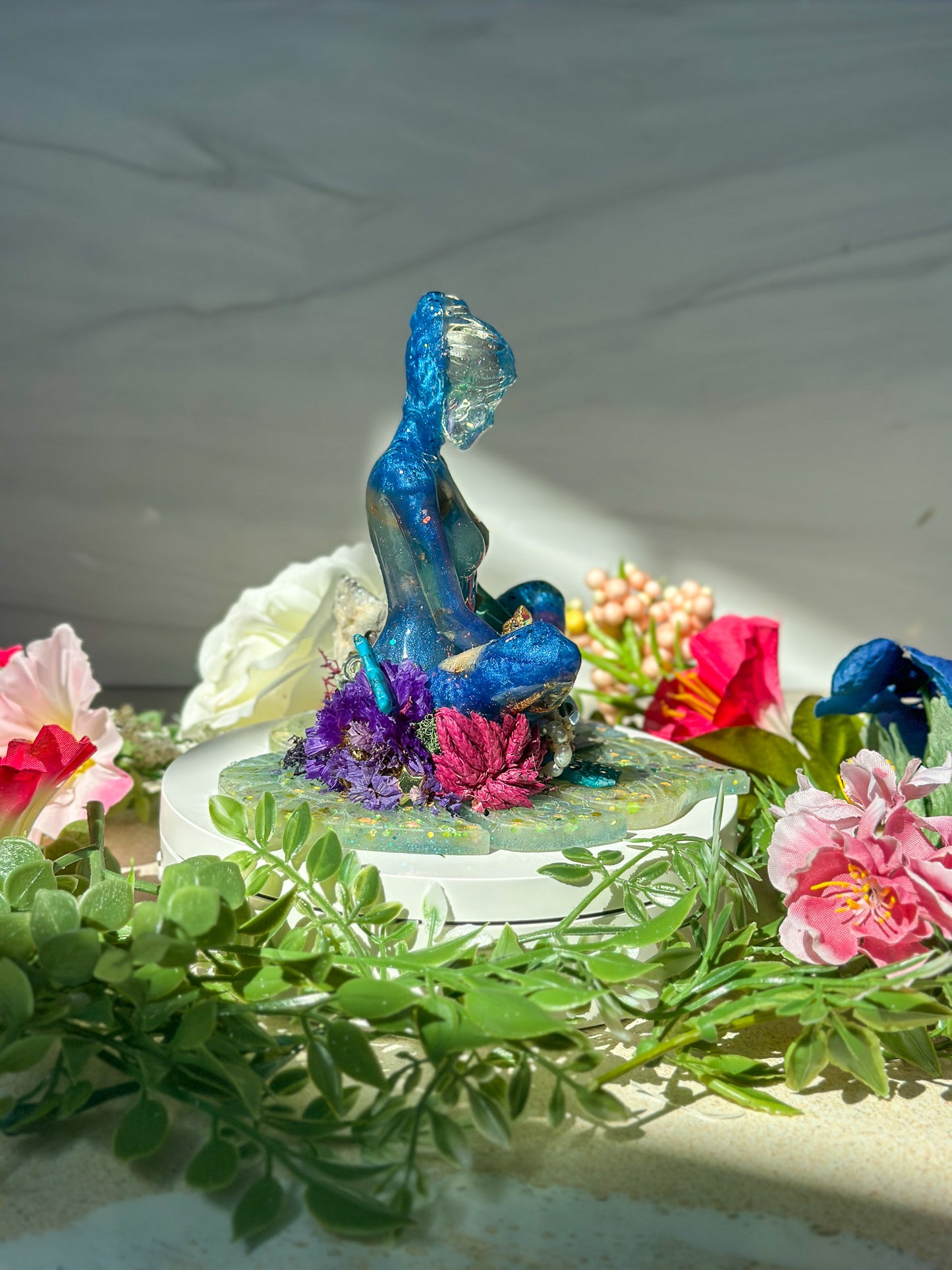 Decorative figurine of a meditating goddess sitting on a resin monstera leaf, surrounded by flowers and greenery.
