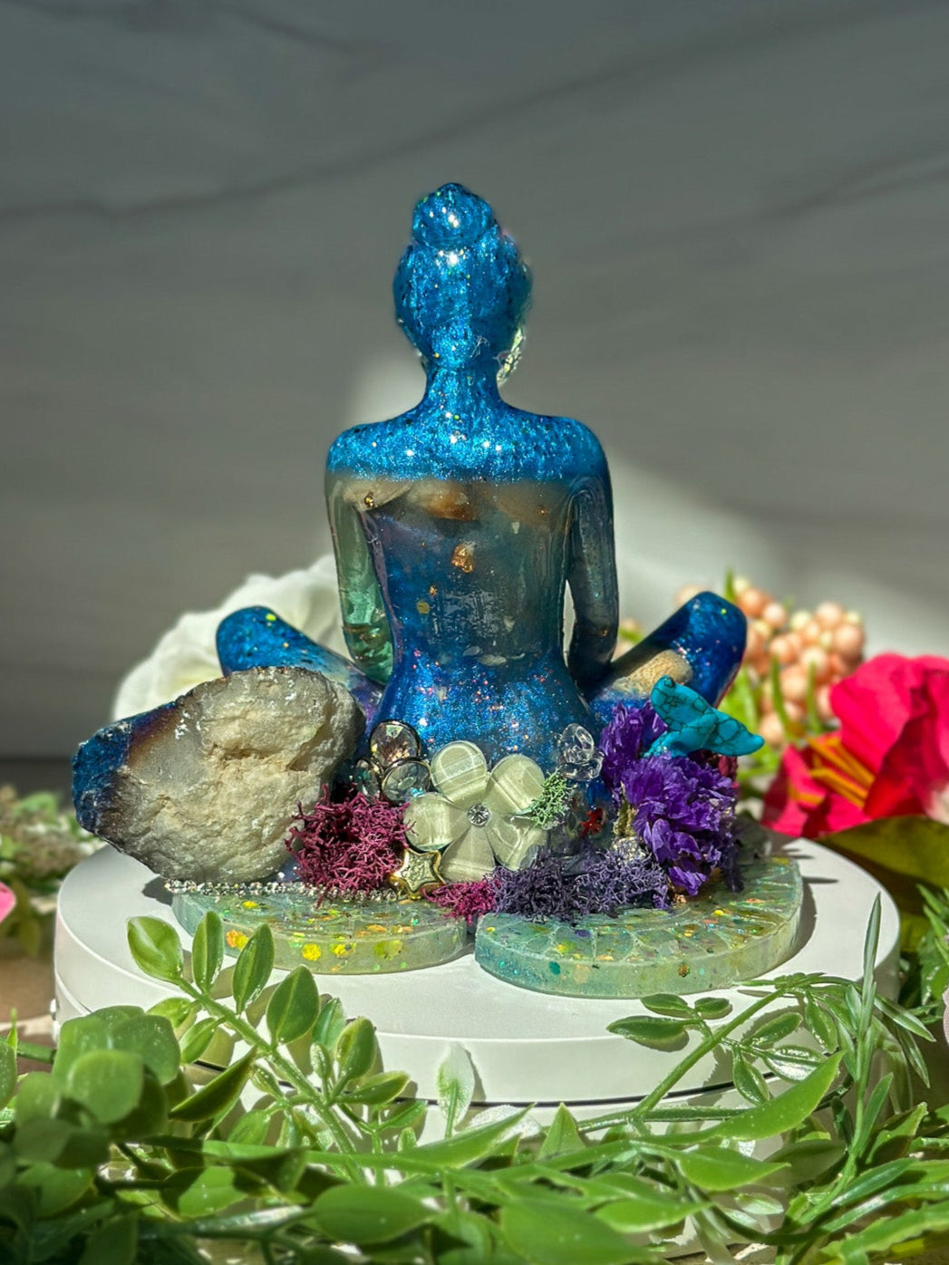 Decorative setup with a blue meditating woman's back, flowers, and greenery on a white surface.