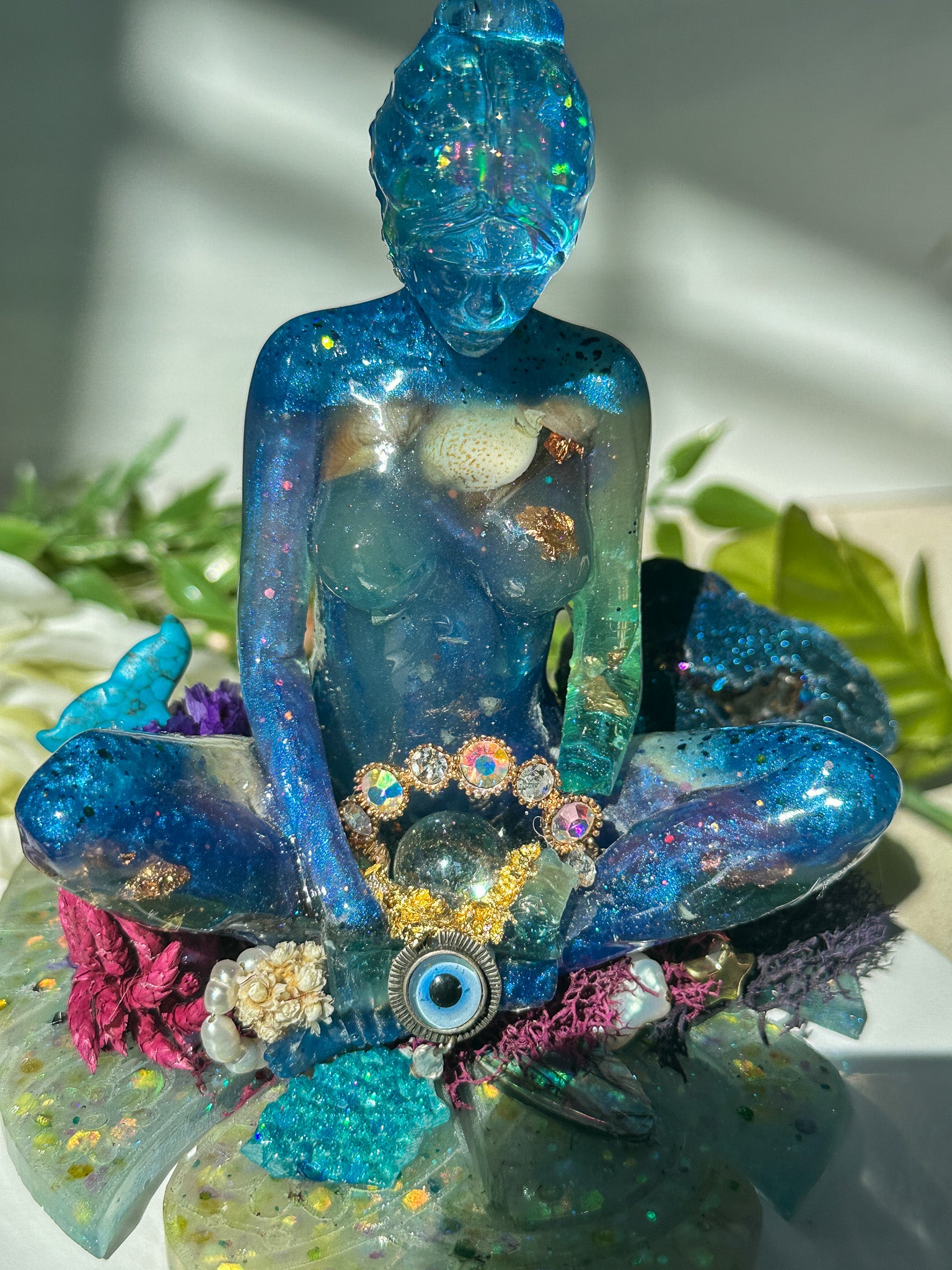 Decorative figurine of a blue Buddha-like figure with gemstones and flowers on a white surface.