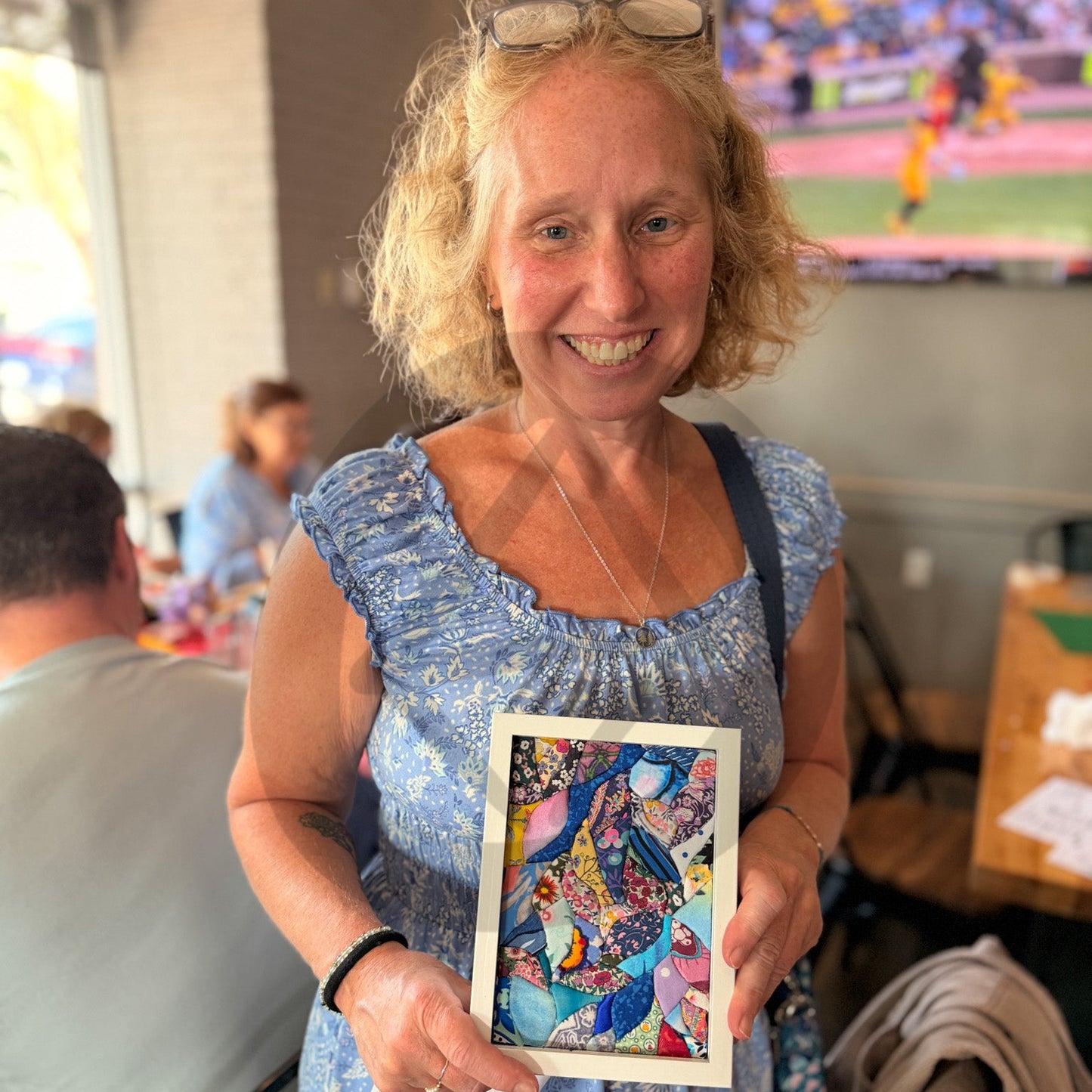 A smiling blonde woman in a blue patterned dress holds up a small framed colorful hand-cut Kinusaiga artwork in a restaurant, with a TV and seated people blurred in the background.