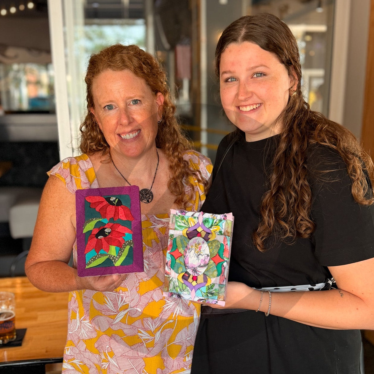 Two women standing indoors and smiling while holding colorful Advanced Kinusaiga artwork pieces. The woman on the left wears a yellow and pink floral dress, and the woman on the right wears a black dress. Both Kinusaiga designs were brought in by these participants.