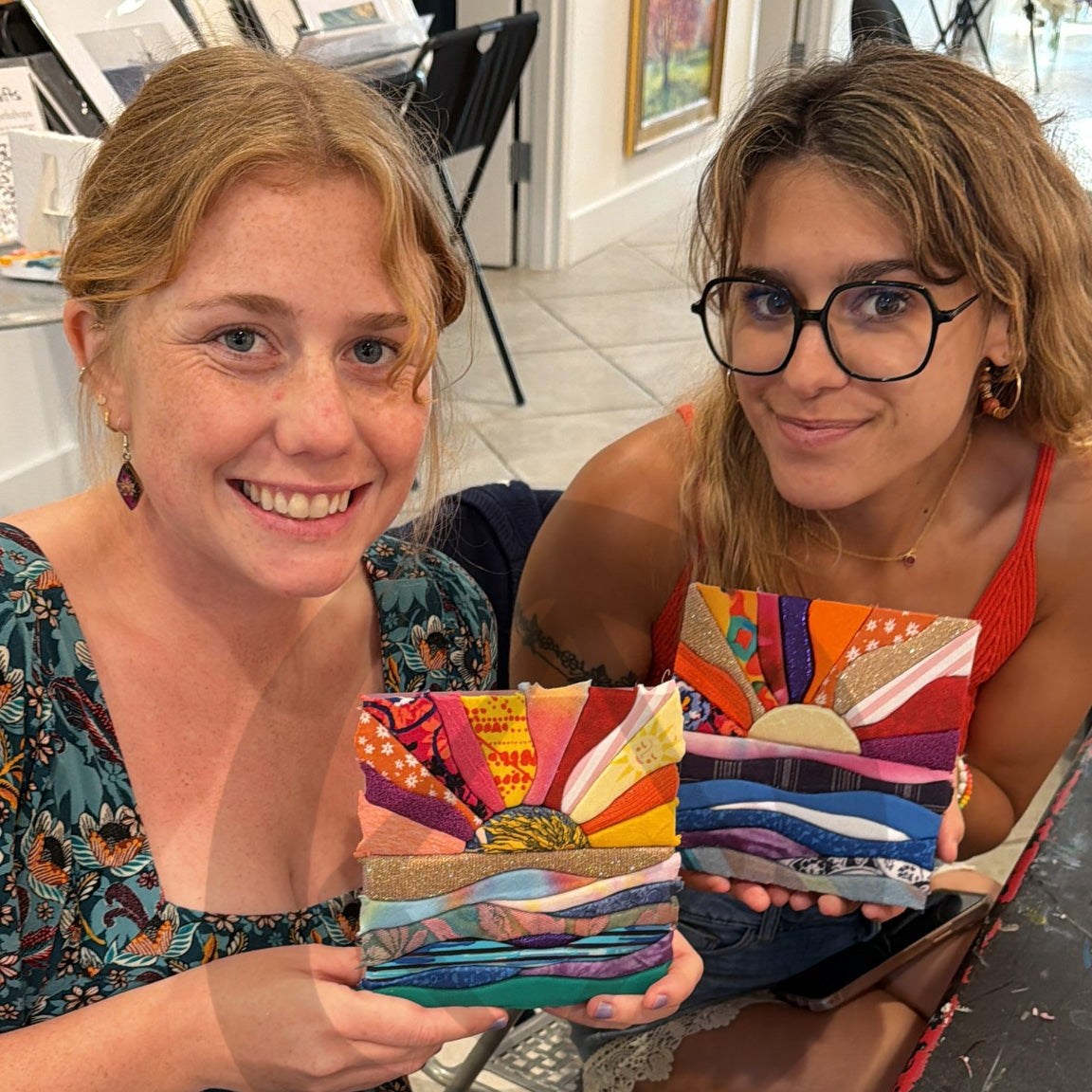 Two women sitting at a craft table holding colorful 6x6 pre-cut sun on waves Kinusaiga fabric pieces in an art studio setting.
