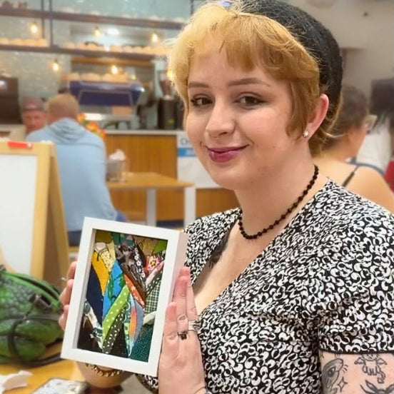A woman with blonde hair and tattoos holds up a square framed 4x6 Kinusaiga, with a colorful abstract design, set in a cafe environment.